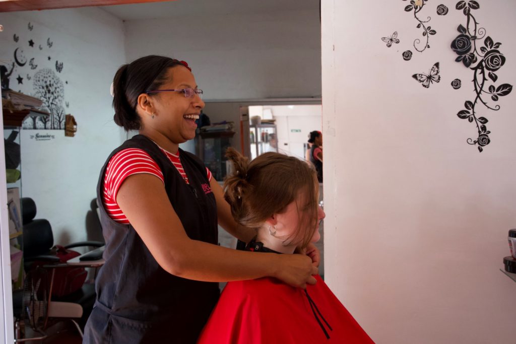 Mary Luz with a client in her hair salon. The client sitting in the salon chair and Marry standing behind her putting on the cape.
