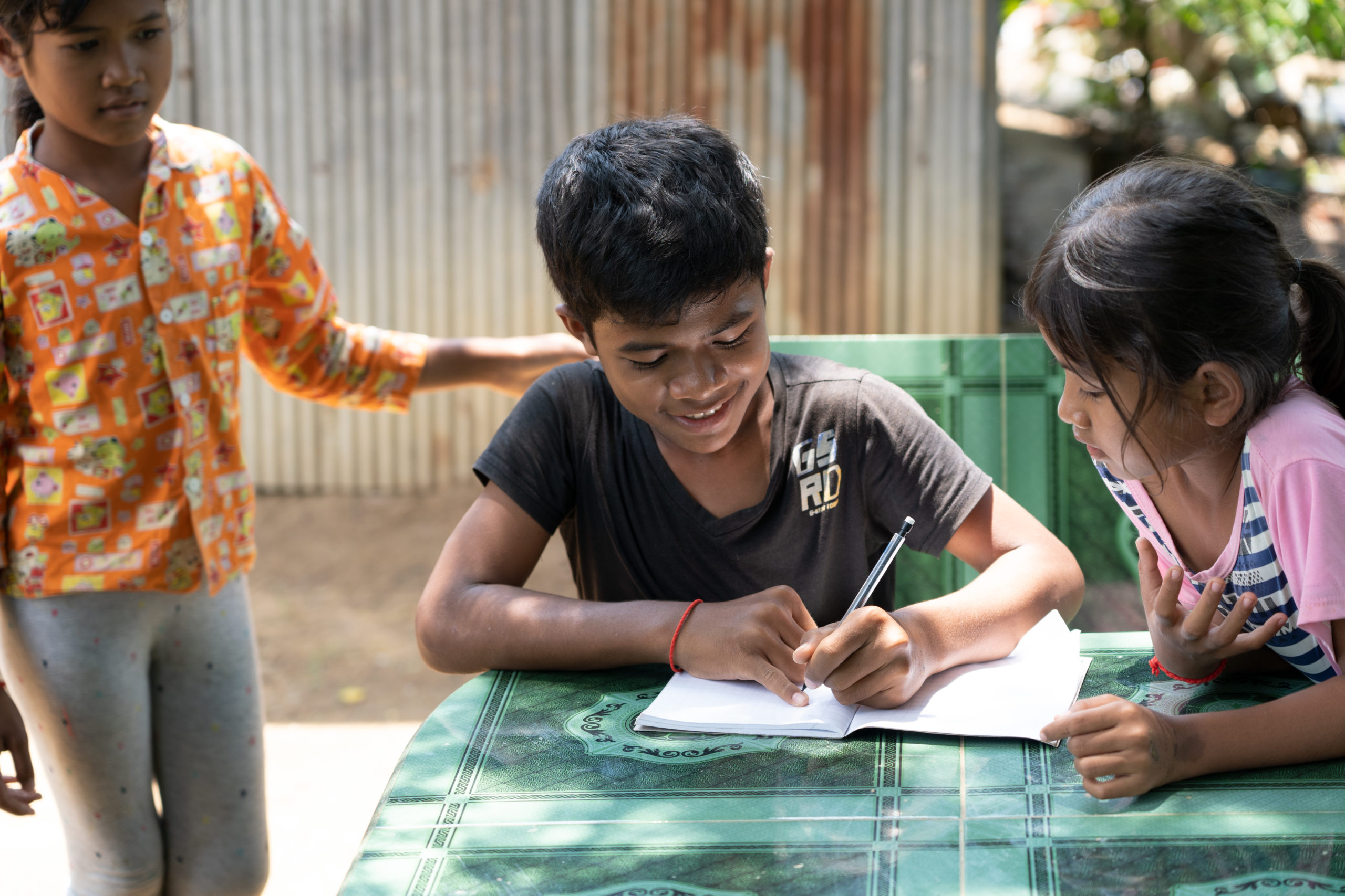a boy in cambodia returns from an orphanage to stay with relatives. he sits at a table with his relatives and writes in a notebook.