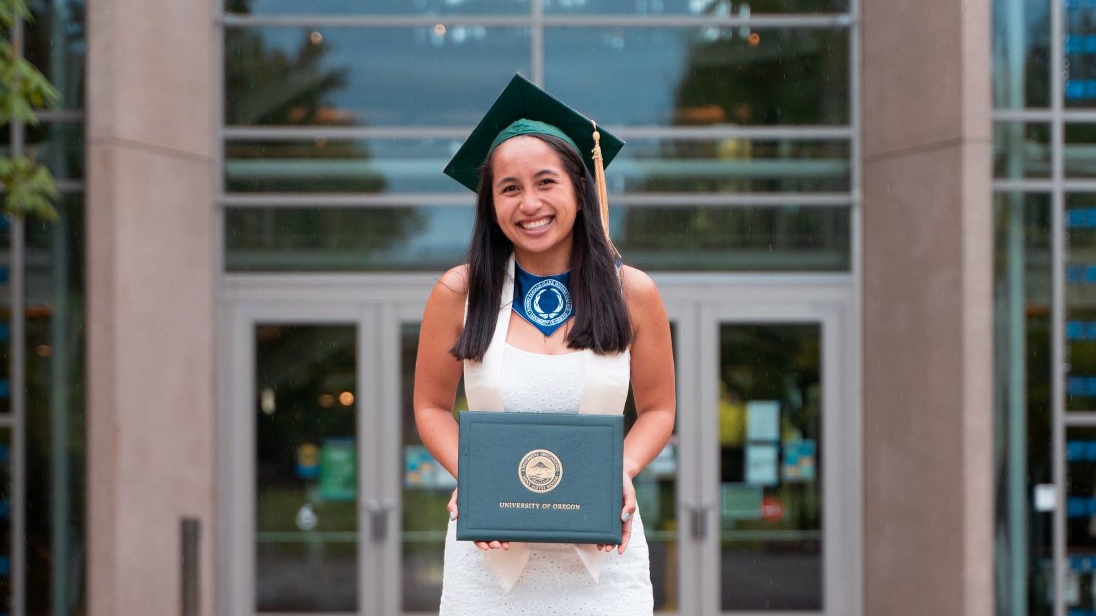 smiling girl wearing green graduation cap and white dress holding diploma in front of big yellow O