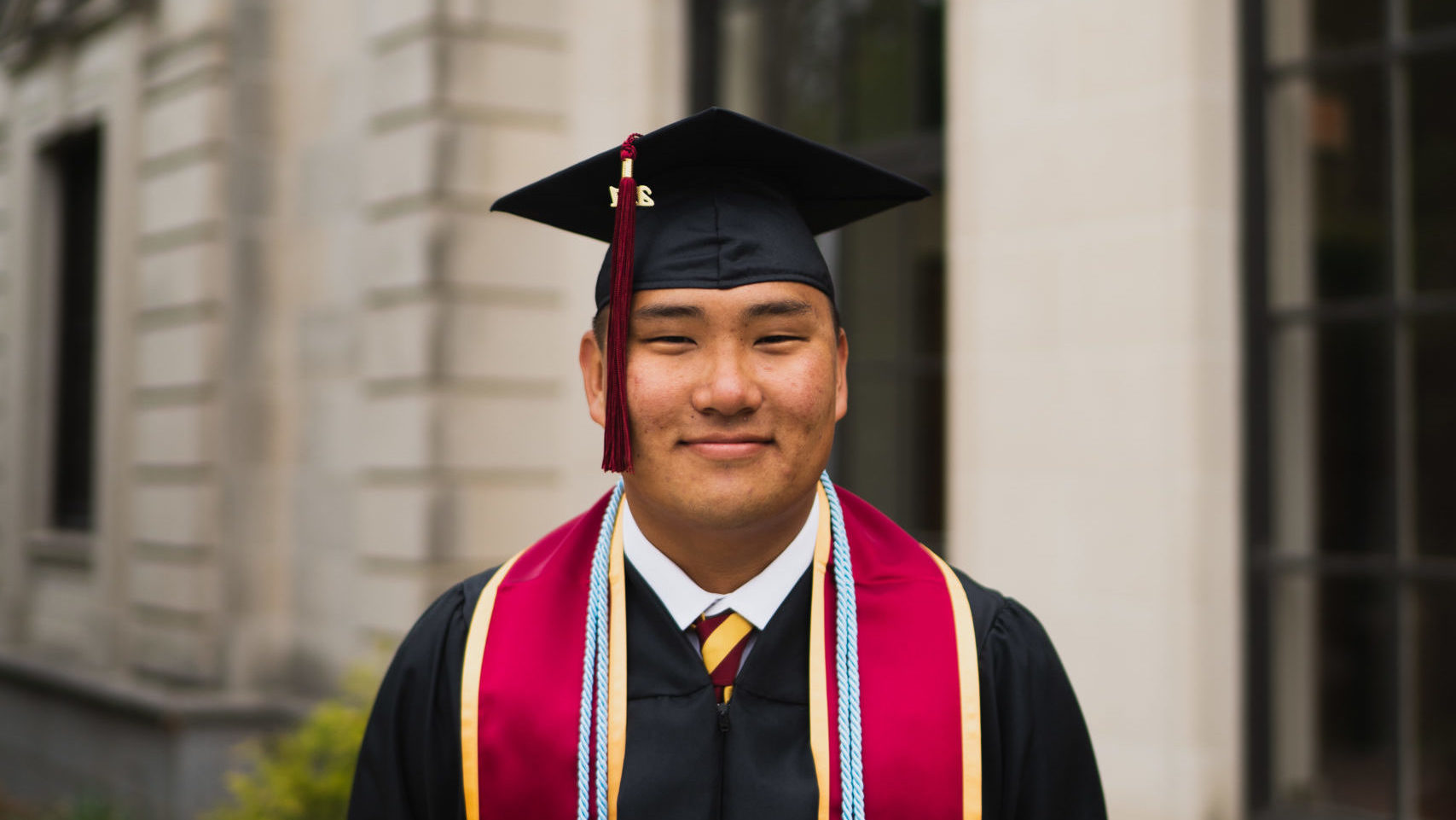 smiling man wearing black graduation cap and gown and burgundy sash