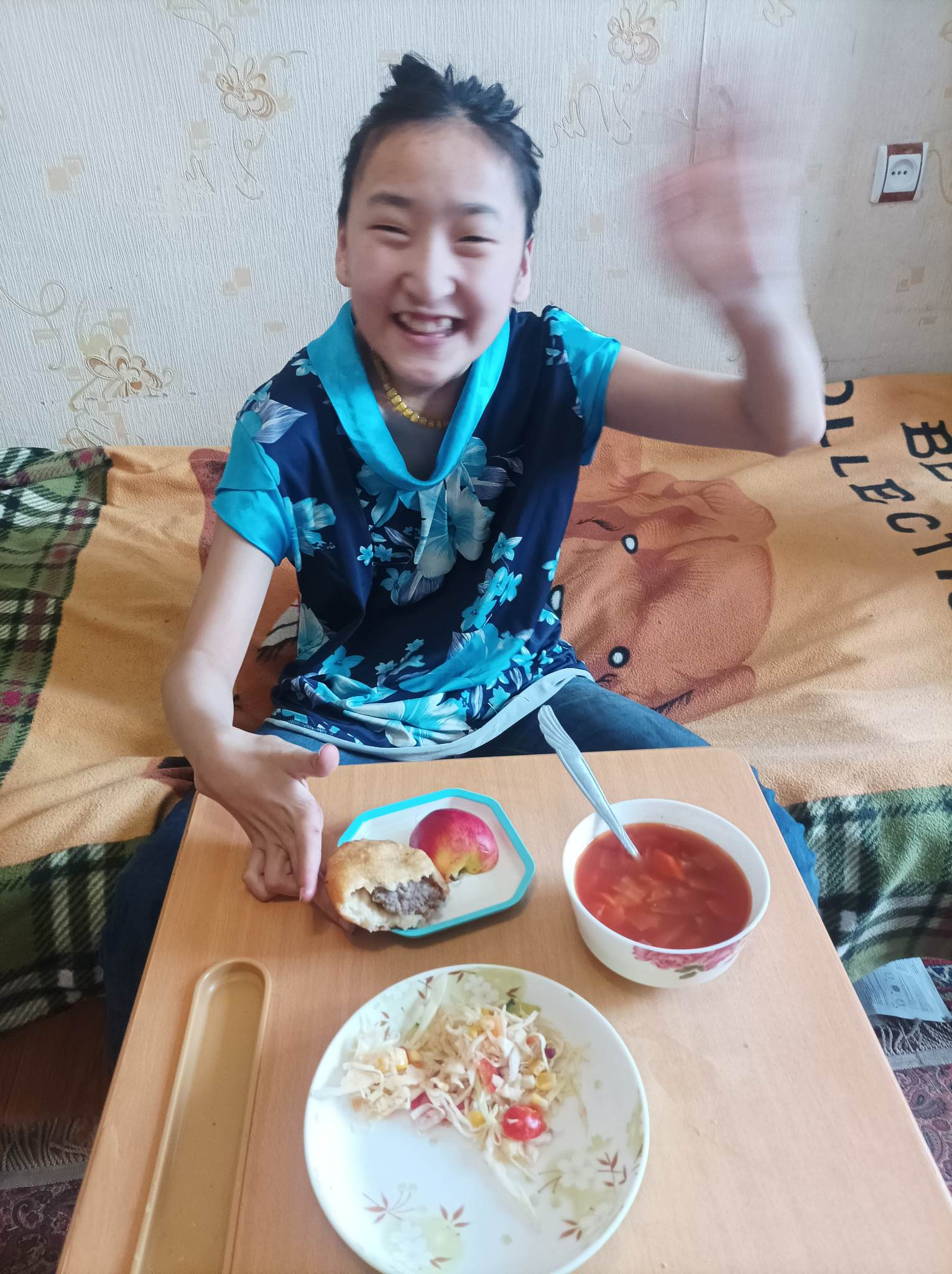 Girl smiles and waves to camera with school lunch on table in front of her