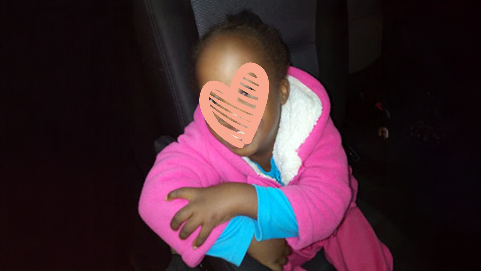 a little girl in a pink sweatshirt smiles with a heart over her face. she is waiting to be adopted.