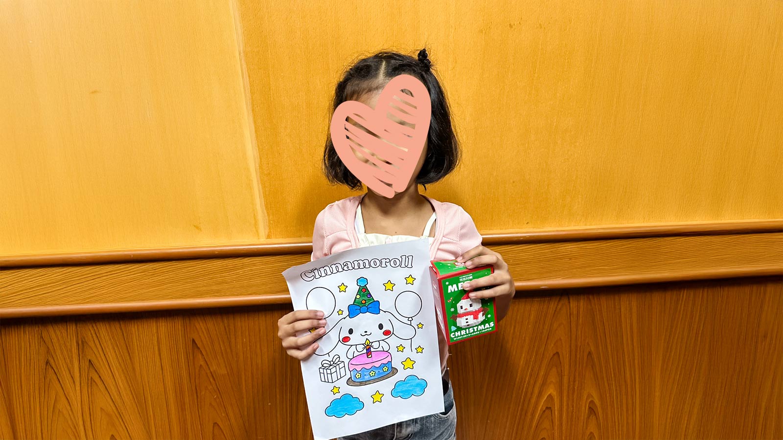 a little girl with a heart over her face stands against a wall and holds up a coloring sheet she colored
