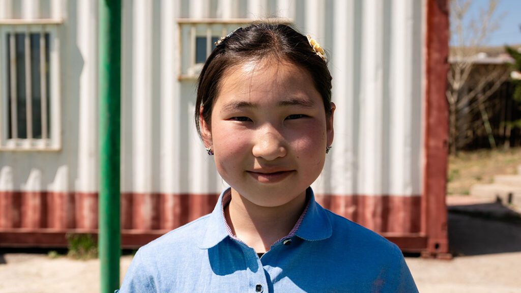 A girl smiles slightly at the camera in front of a school