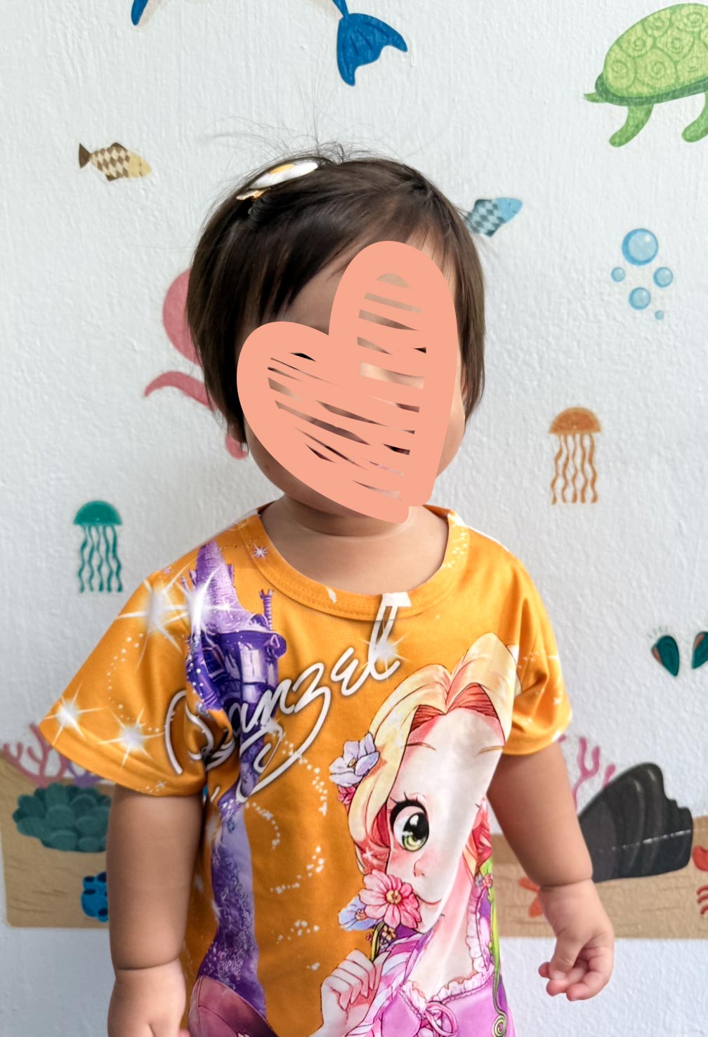 A little girl stands against a wall and is wearing a princess dress with a heart over her face.