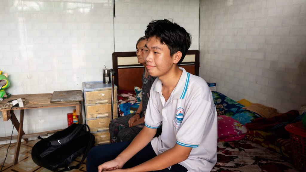 grandson and grandmother sit on the edge of the grandma's bed that is located in the living room