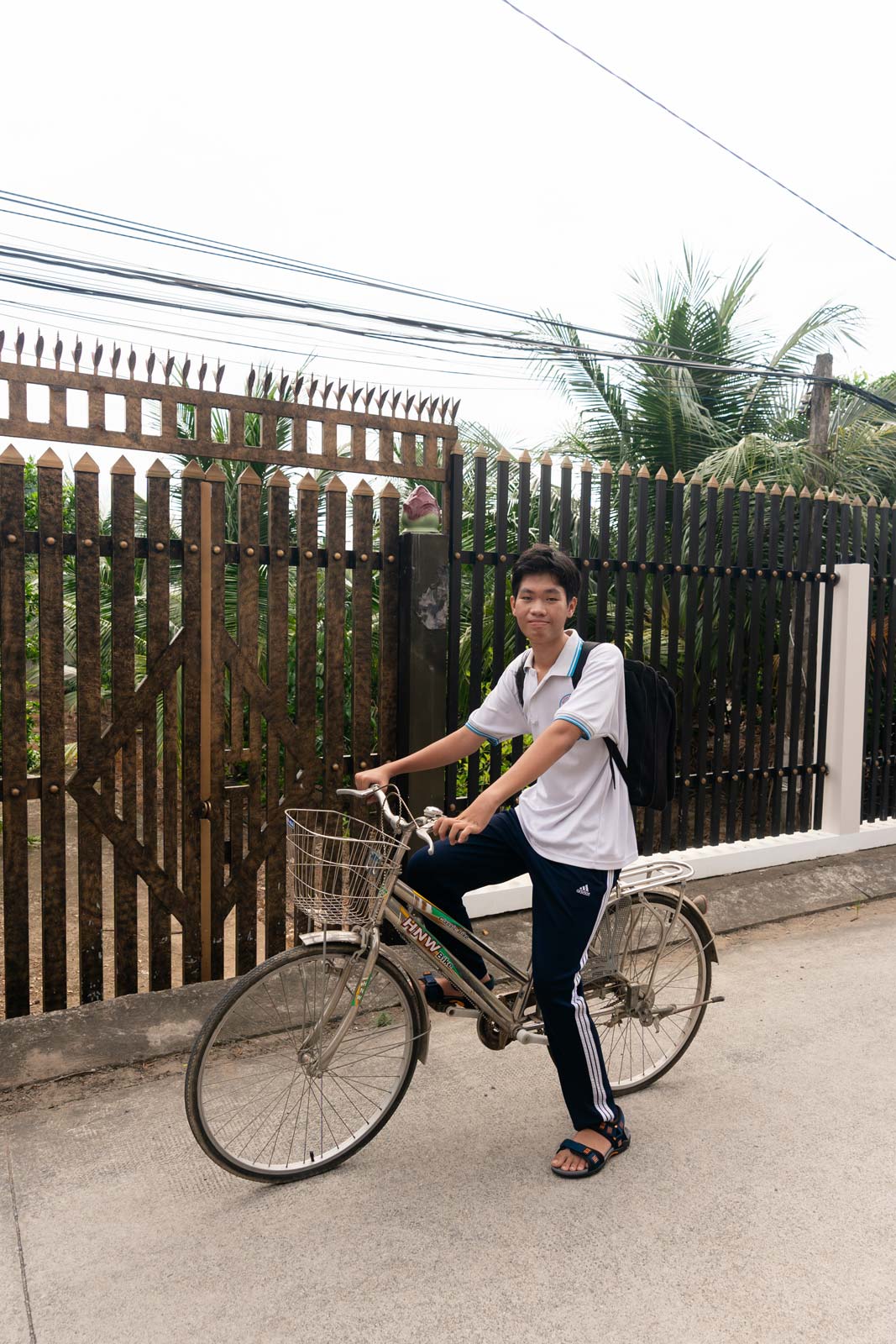 teenage boy poses, half smiling on his bike in the street across from him house, with a fence and palm trees behind him