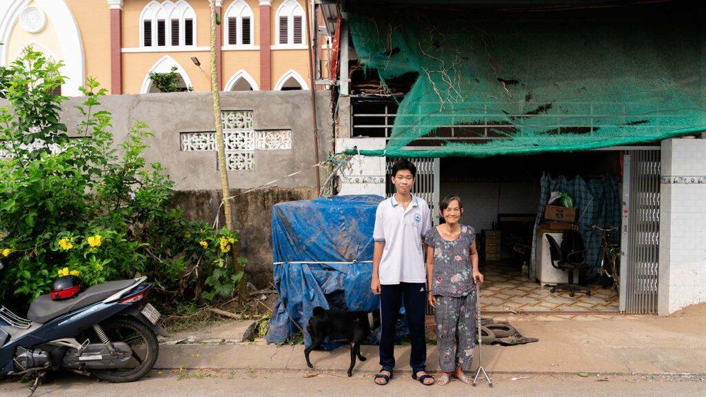 a grandson and grandmother with a cane stand next to each other, and their dog outside of their home in Vietnam