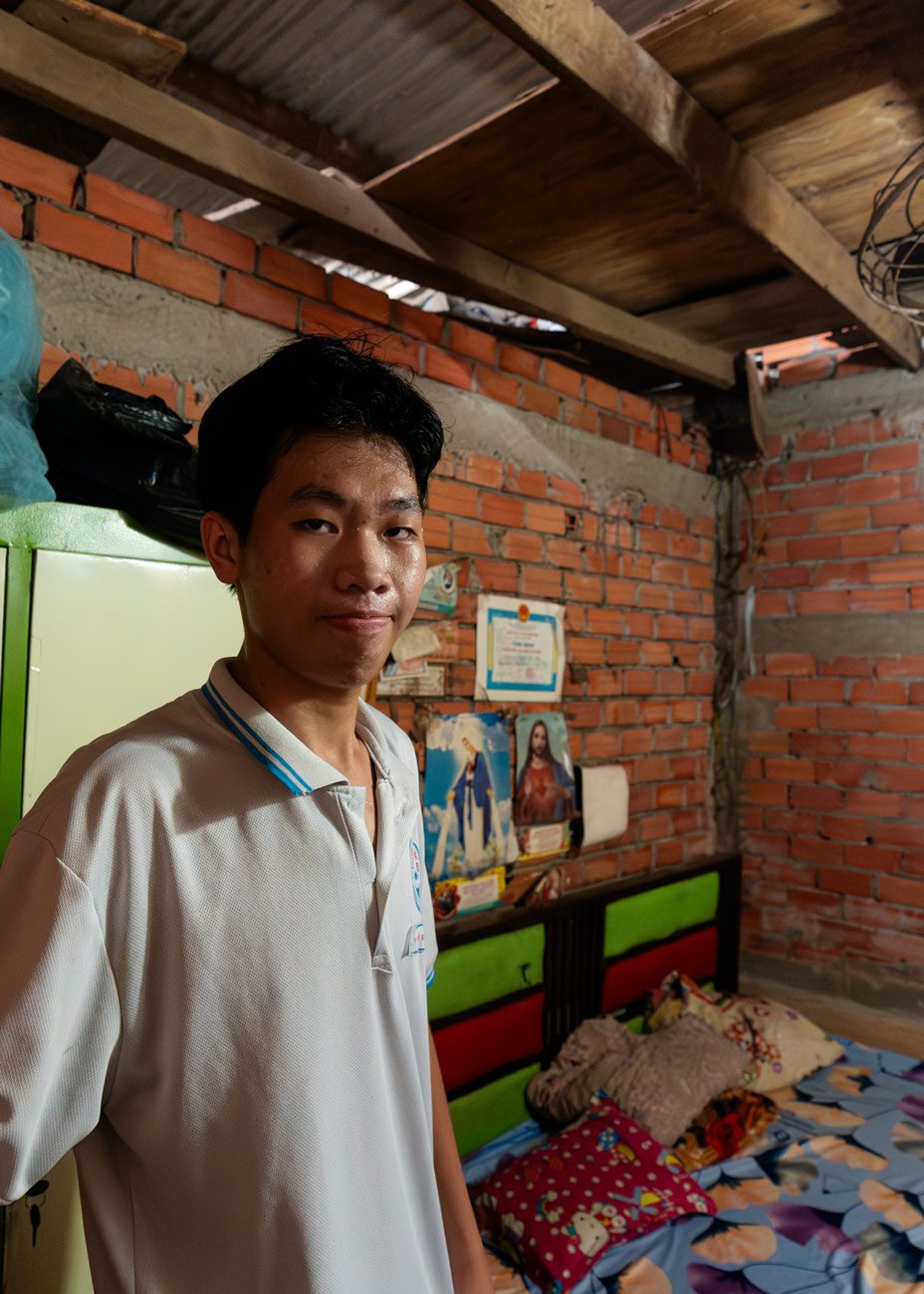 close up of a teenage boy standing in his room, in front of his bed, staring at the camera