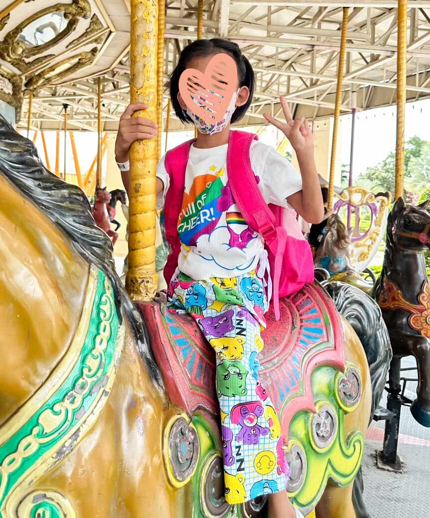 a little girl with a heart over her face sits on a carousel with a colorful outfit. She needs an adoptive family.