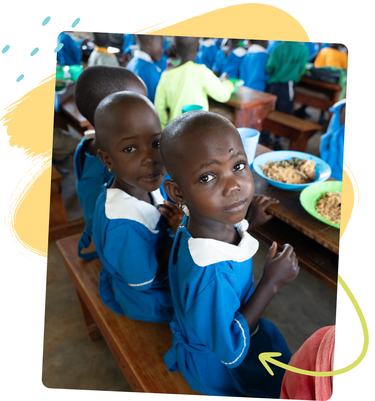Photo of several children in blue school uniforms sitting at a wooden table with full plates of food in front of them. 