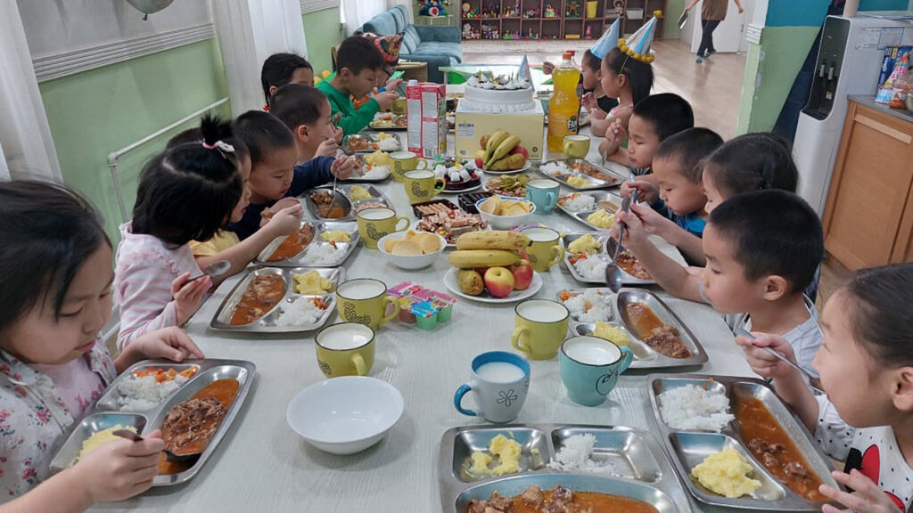 Photograph of a long table full of children eating from their plates. The table is overflowing with healthy foods. 