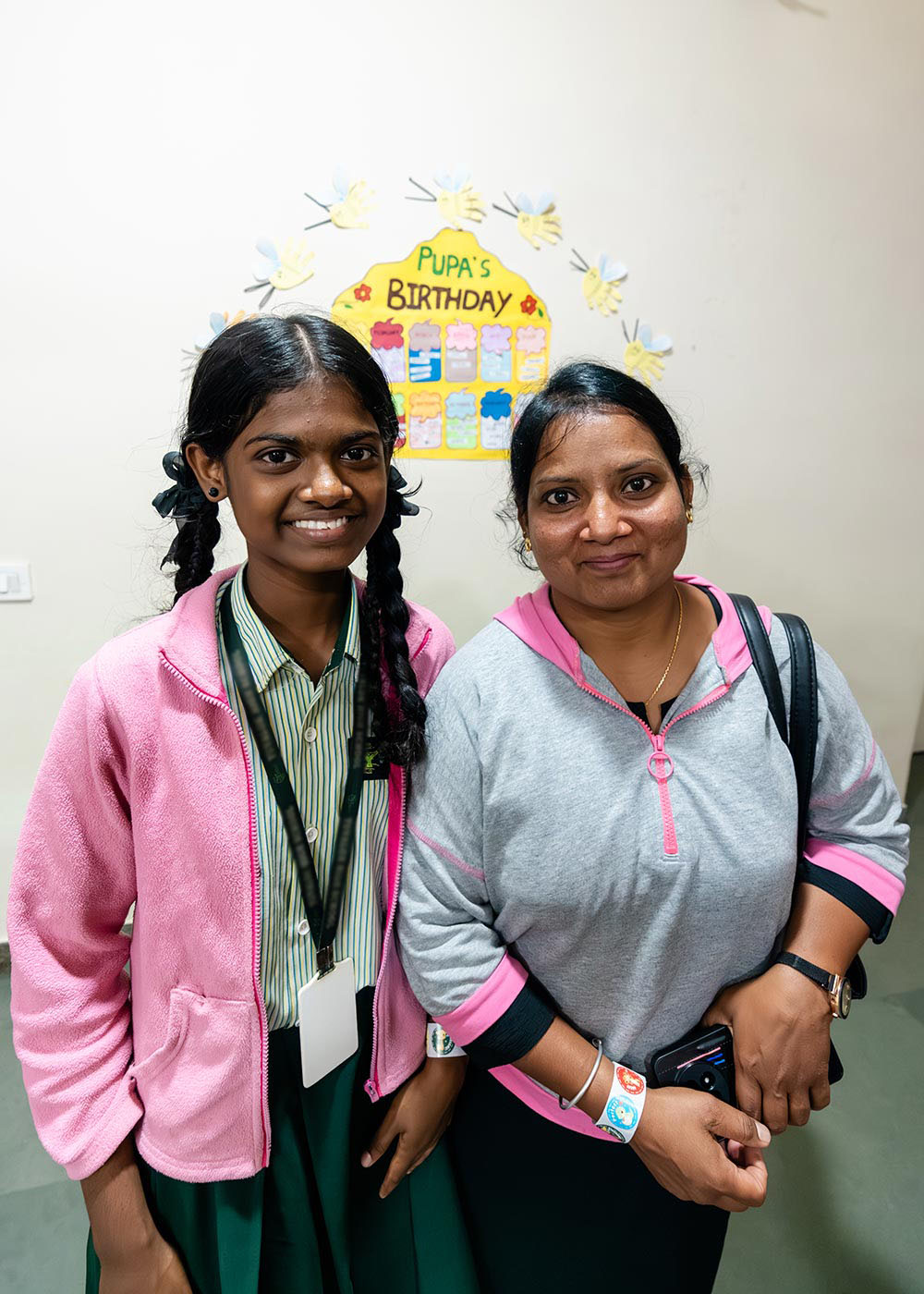 a sponsored girl in a school uniform and pink jacket smiles next to her mom in a preschool classroom