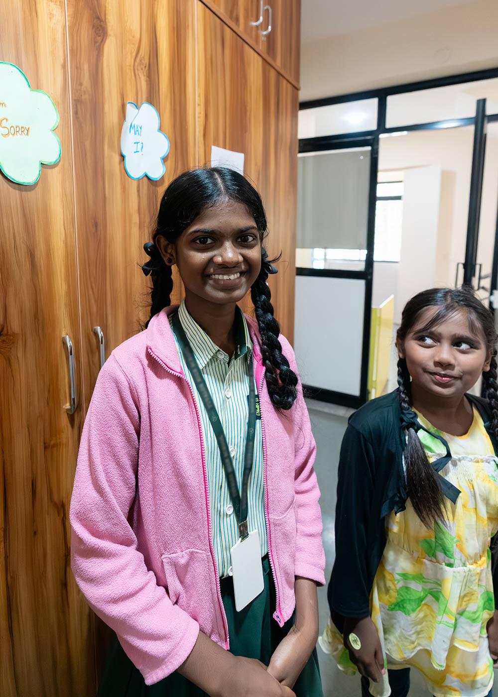 a sponsored girl in a school uniform and pink jacket smiles in a preschool classroom, with younger students