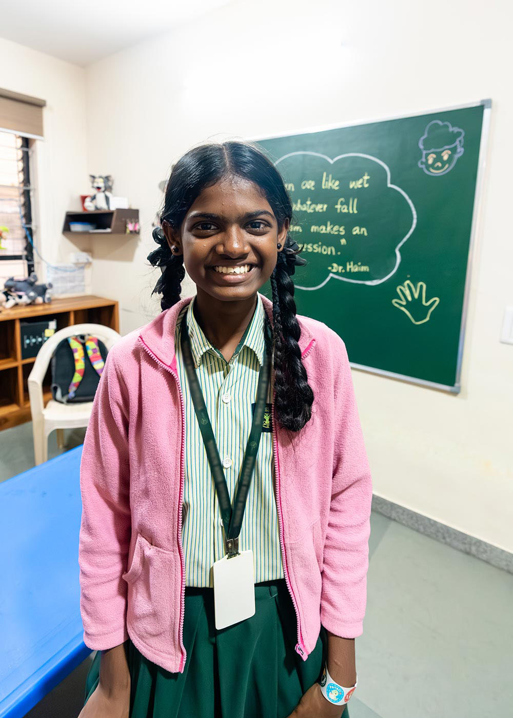 a sponsored girl in a school uniform and pink jacket smiles in a preschool classroom