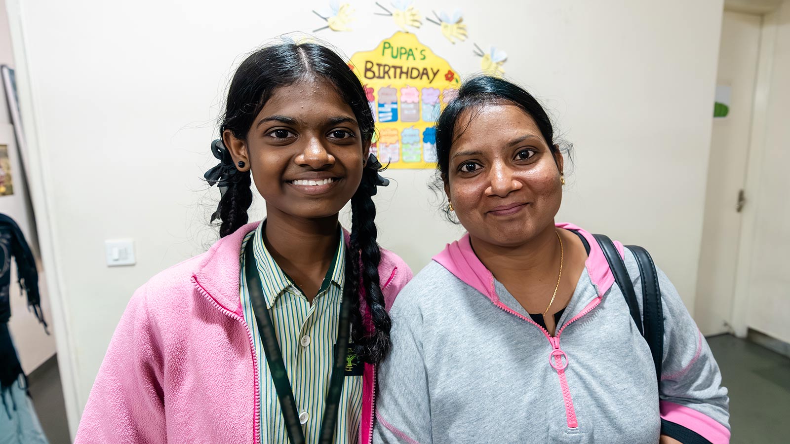 a sponsored girl in a school uniform and pink jacket smiles next to her mom in a preschool classroom