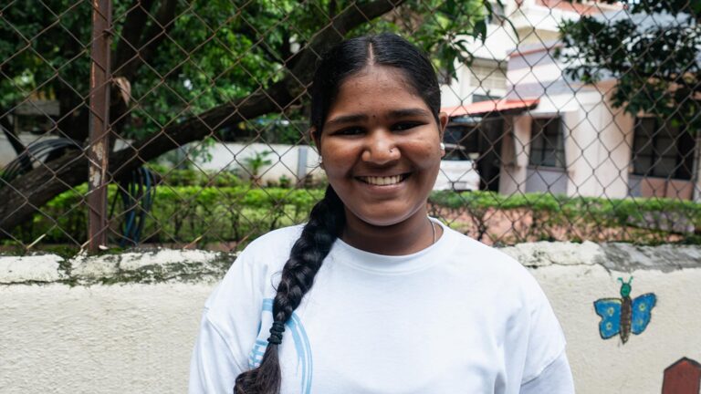 a girl in India stands outside and smiles, wearing a white shirt and a braid