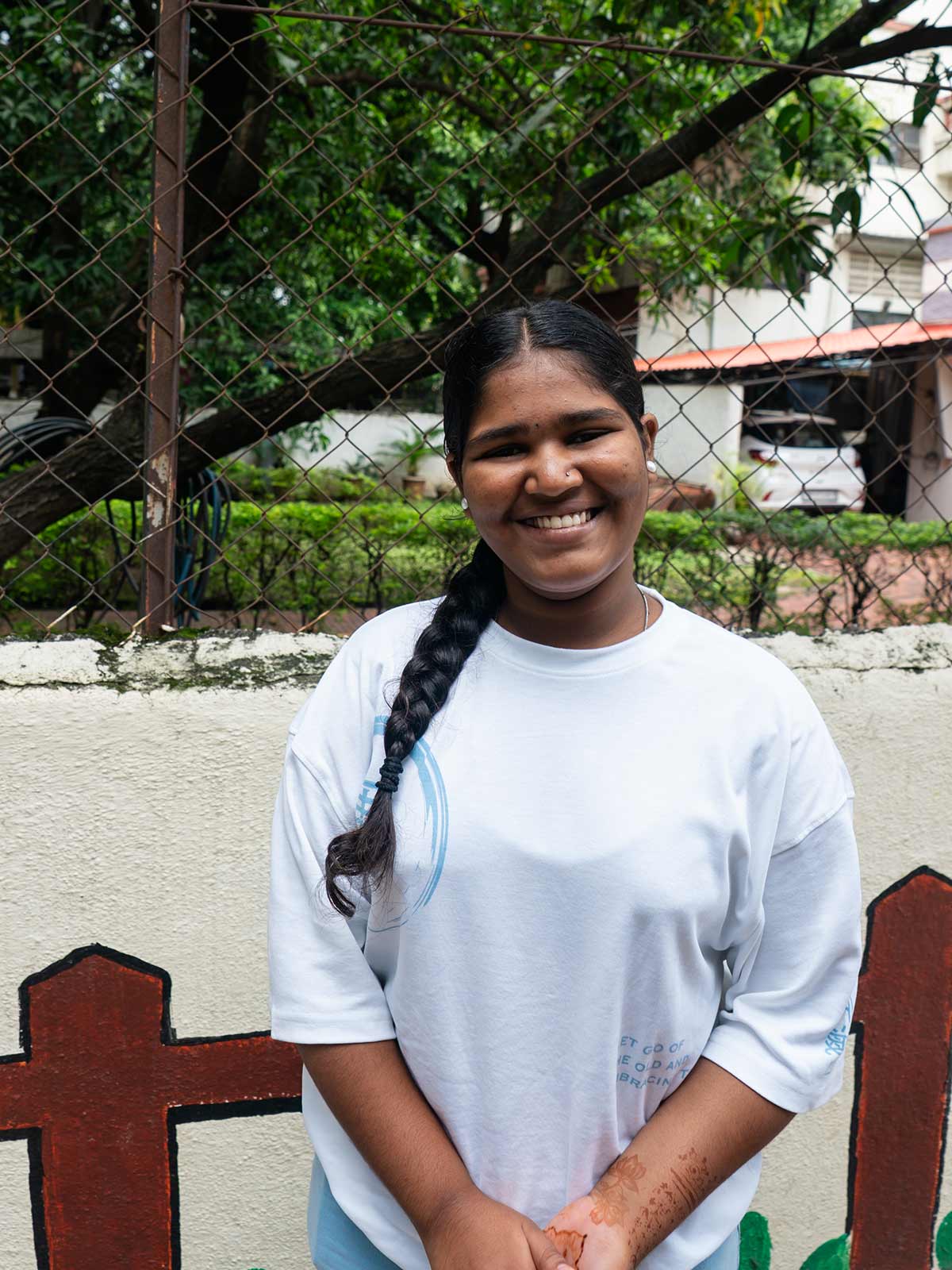 a teenage girl smiles outdoors in India, wearing a white shirt and a long, neat braid.