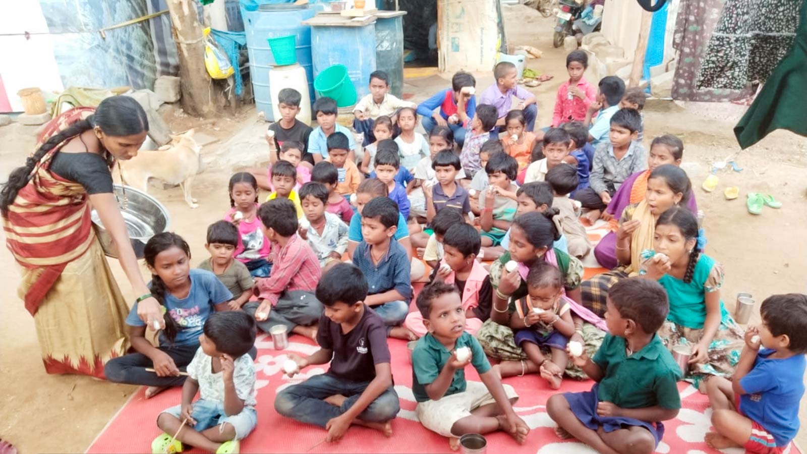 many children sit on a mat in a migrant camp, waiting for a daily meal