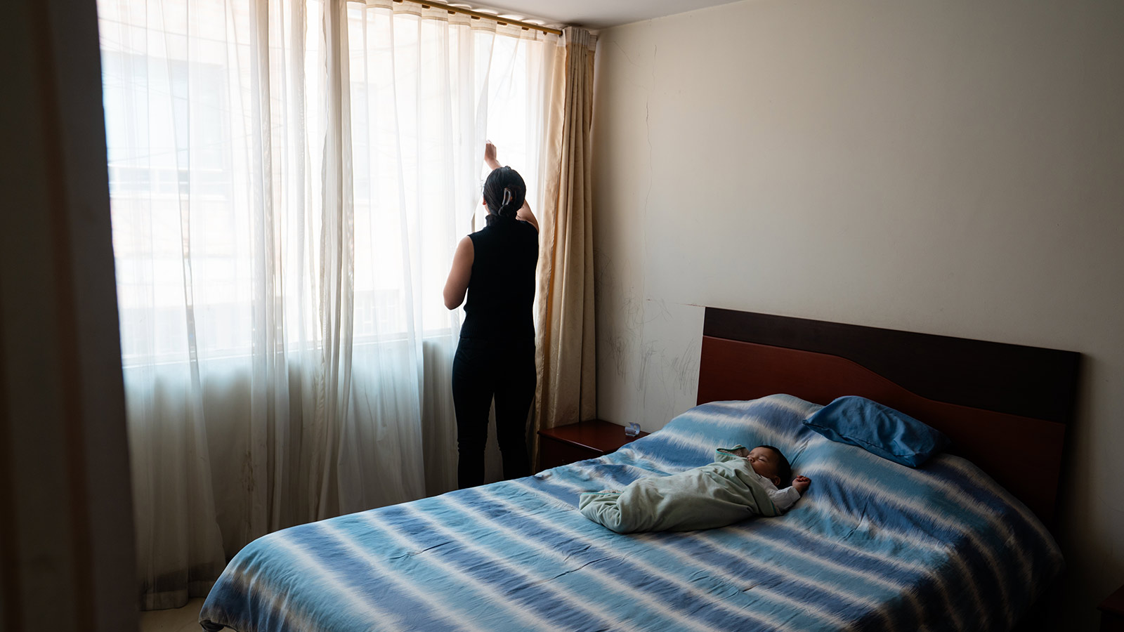 a mother draws the blinds in her bedroom while her son sleeps on the bed