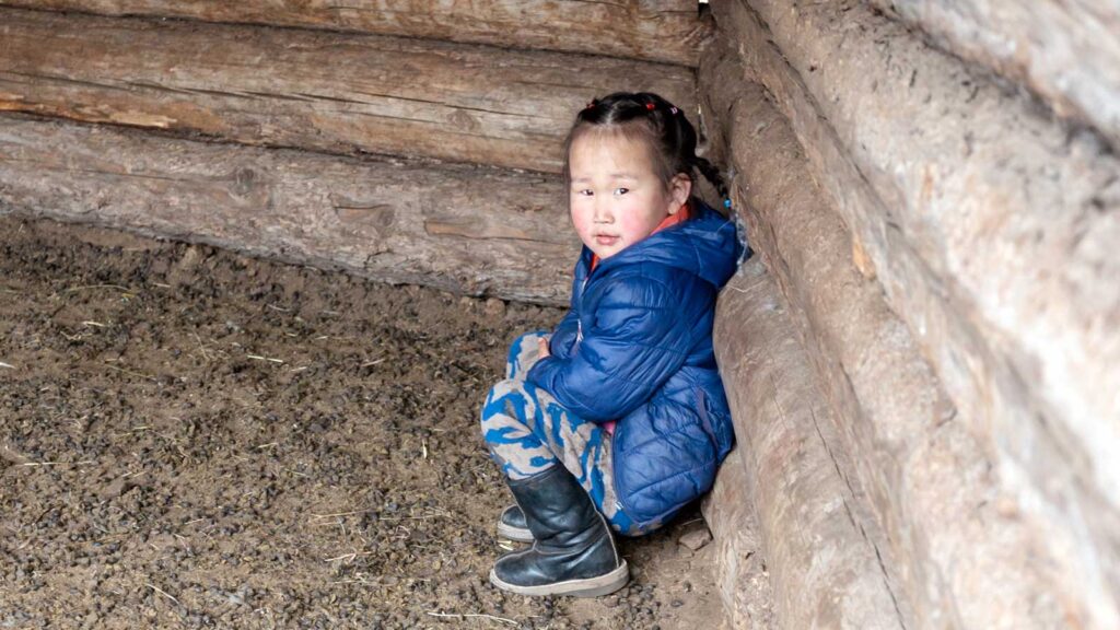 Little girl in Mongolia wearing a blue jacket sitting outside, leaning up against the barn wall