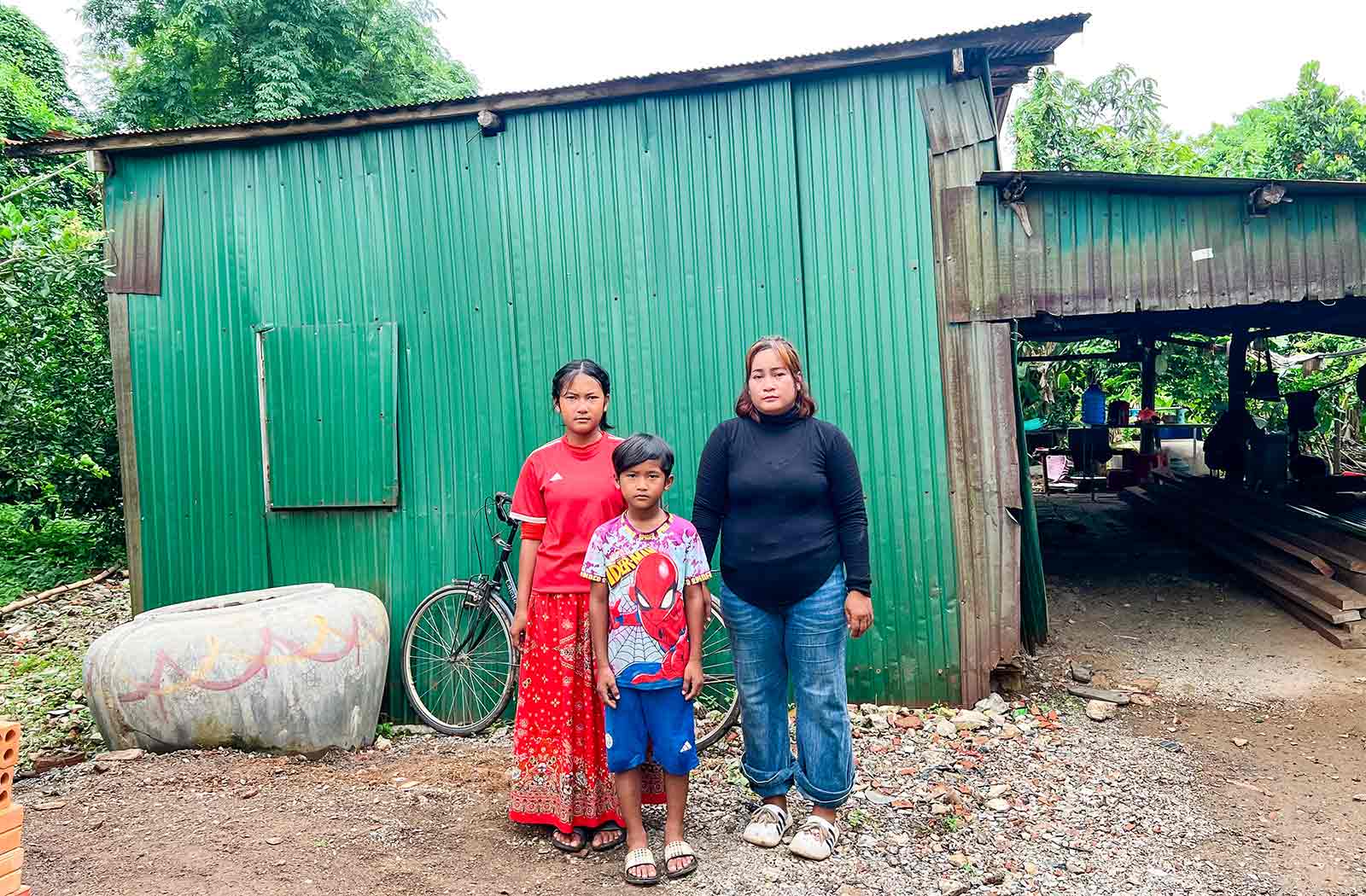 a mother and her two children stand outside of their home — a dilapidated shed in much need of repair