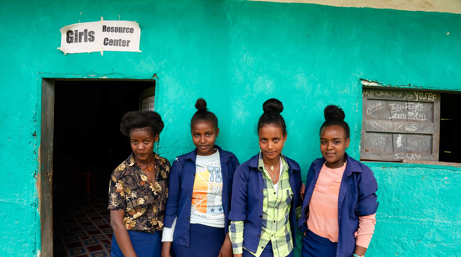 a group of young women stand outside the resource center — a vibrant teal school building