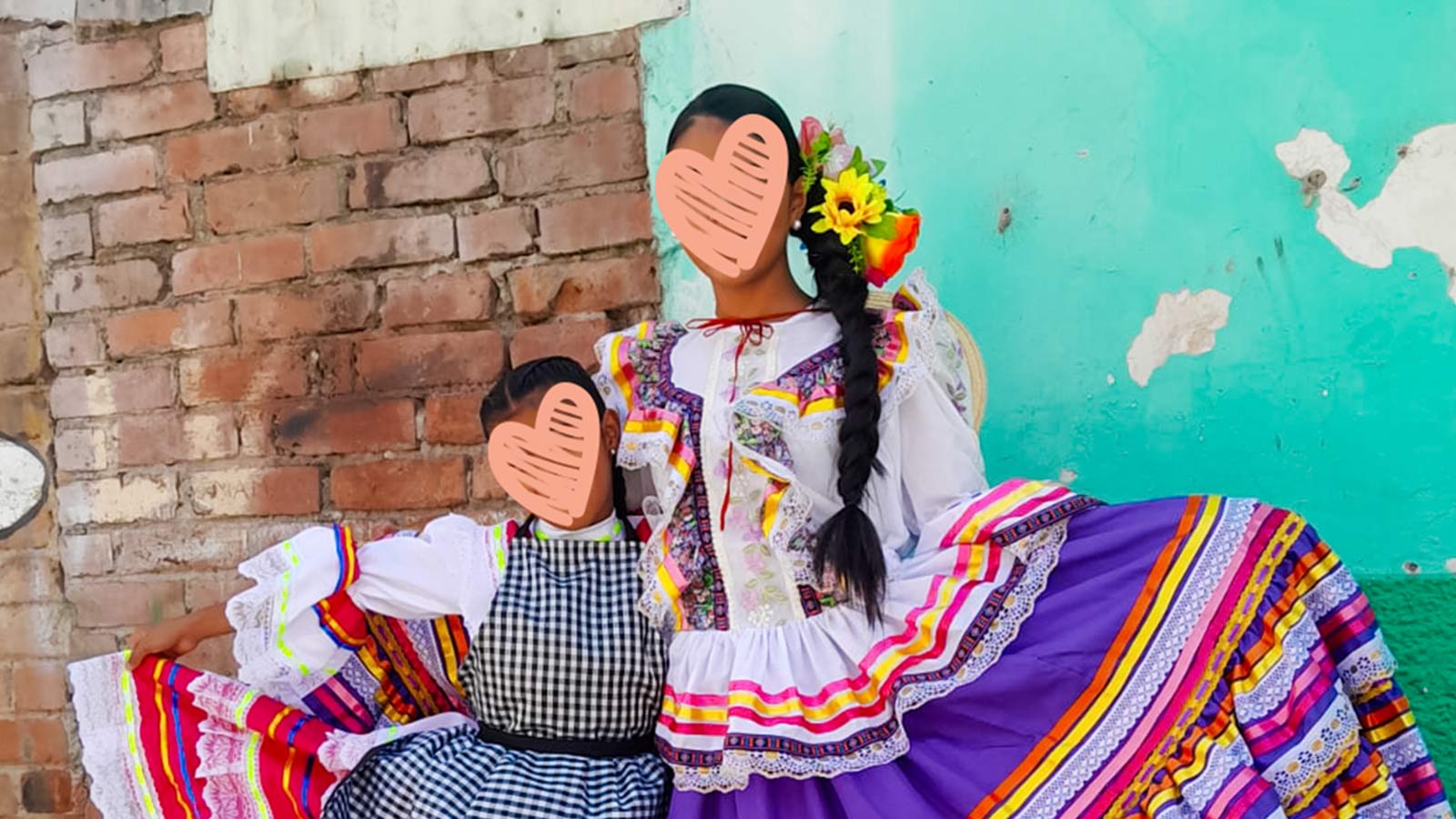two sisters wear beautiful, colorful dress and smile for a photo in front of a teal and brick wall