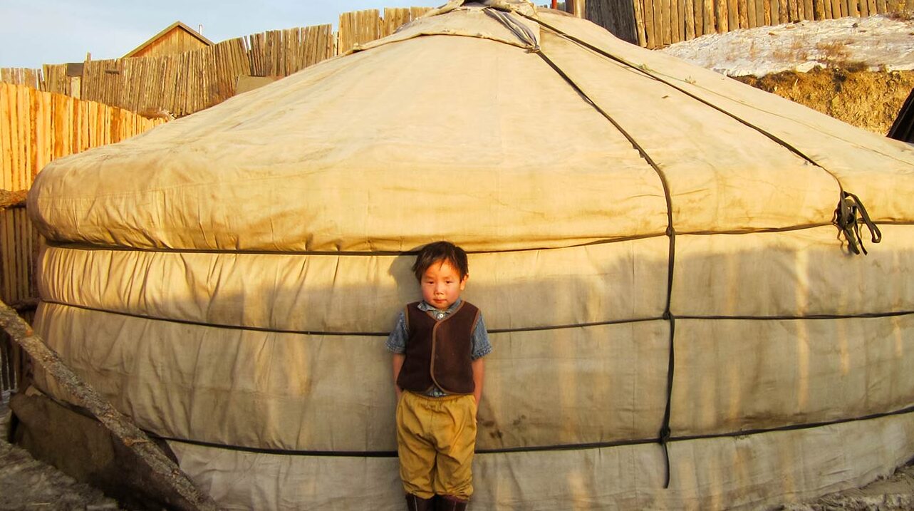 a boy stands outside his ger in Mongolia without a coat in the winter