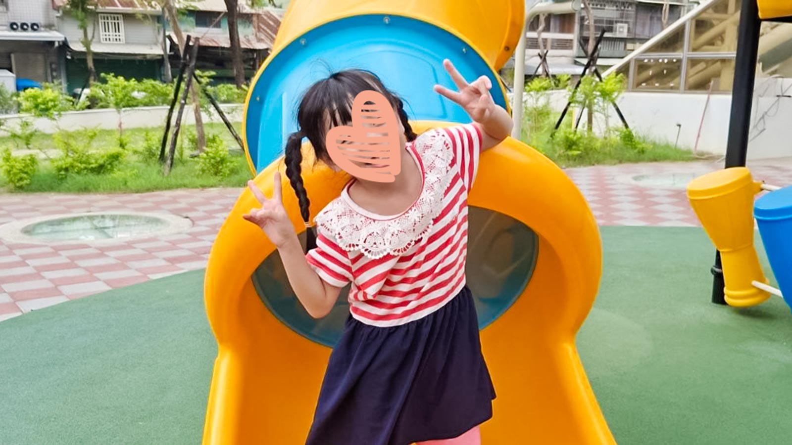 a little girl with a heart over her face stands on a playground slide with peace sign hands