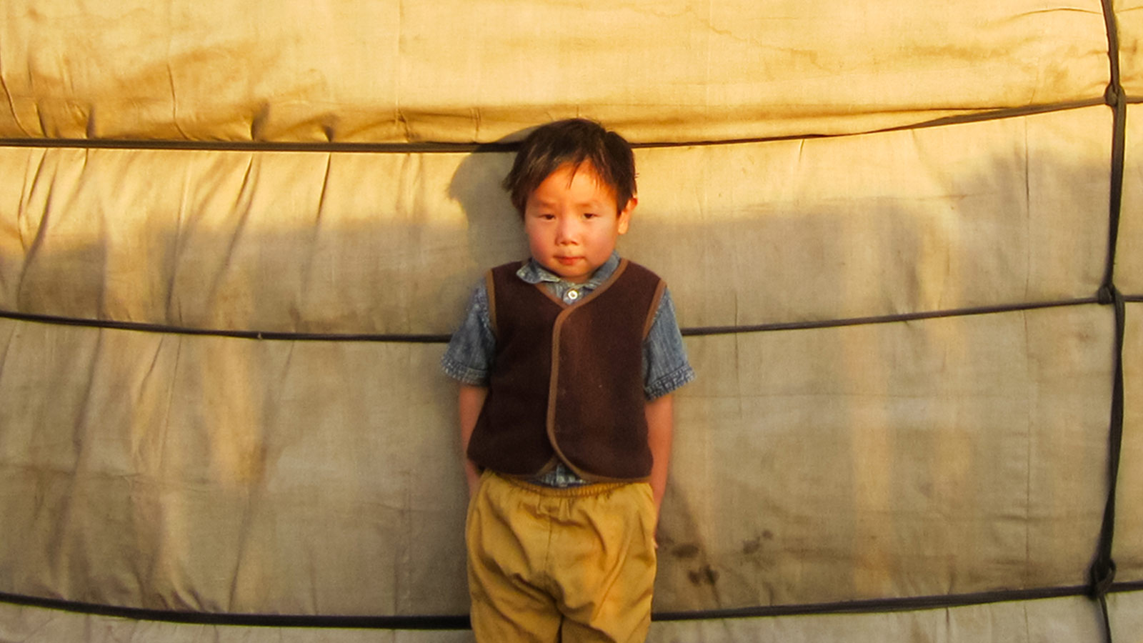 a boy stands outside his ger in Mongolia without a coat in the winter