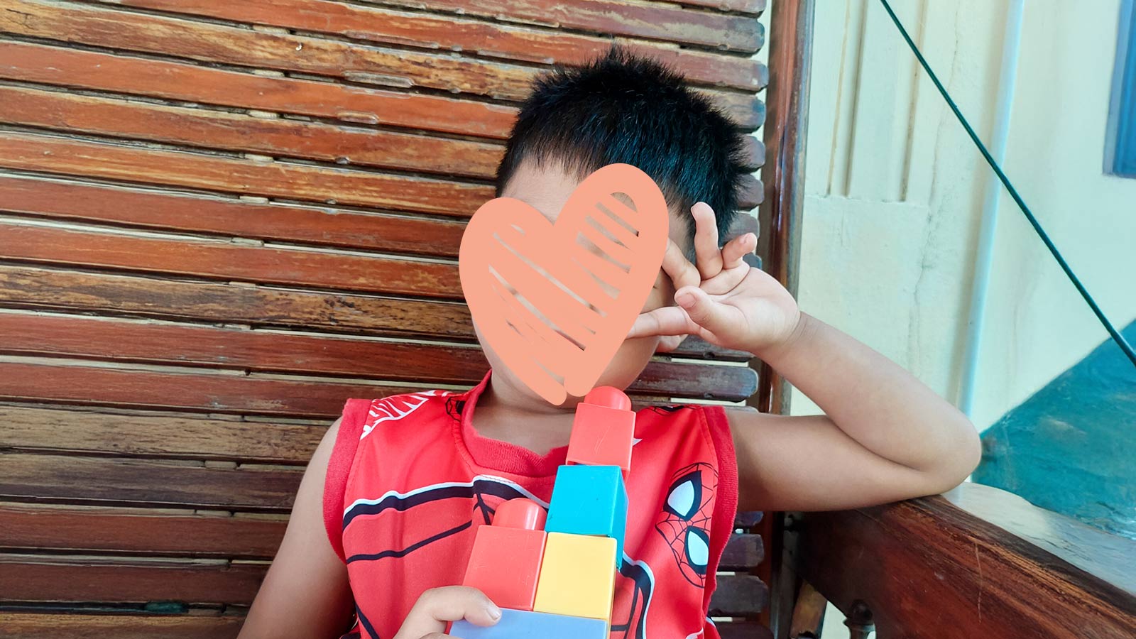 a boy wearing a heart over his face and a spiderman tanktop holds up a friendly wave next to his building blocks