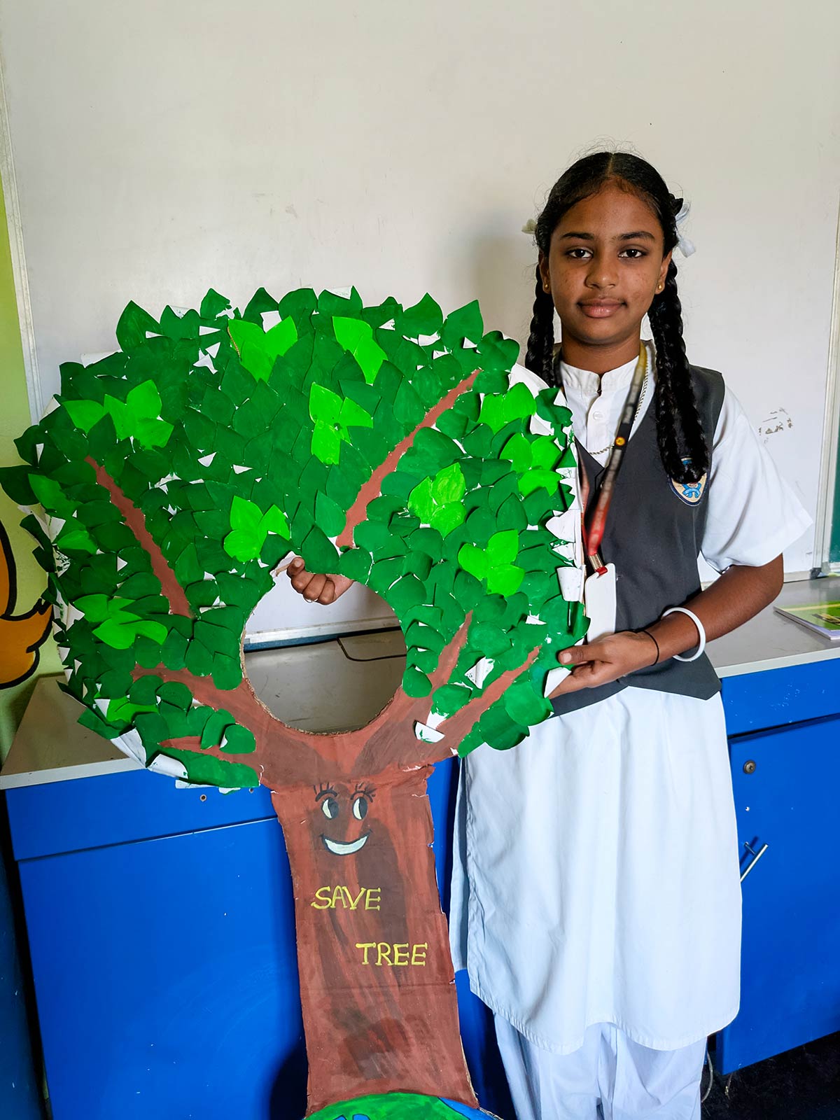 a sponsored girl in India stands next to her art project — a green tree cut out — that she made for school
