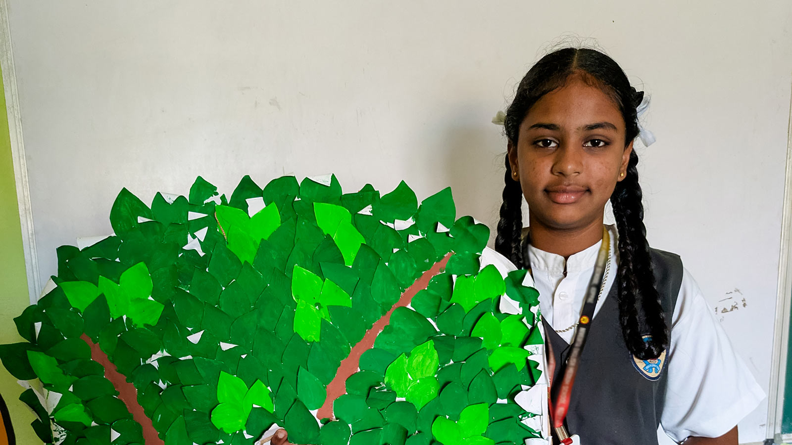 a sponsored girl in India stands next to her art project — a green tree cut out — that she made for school