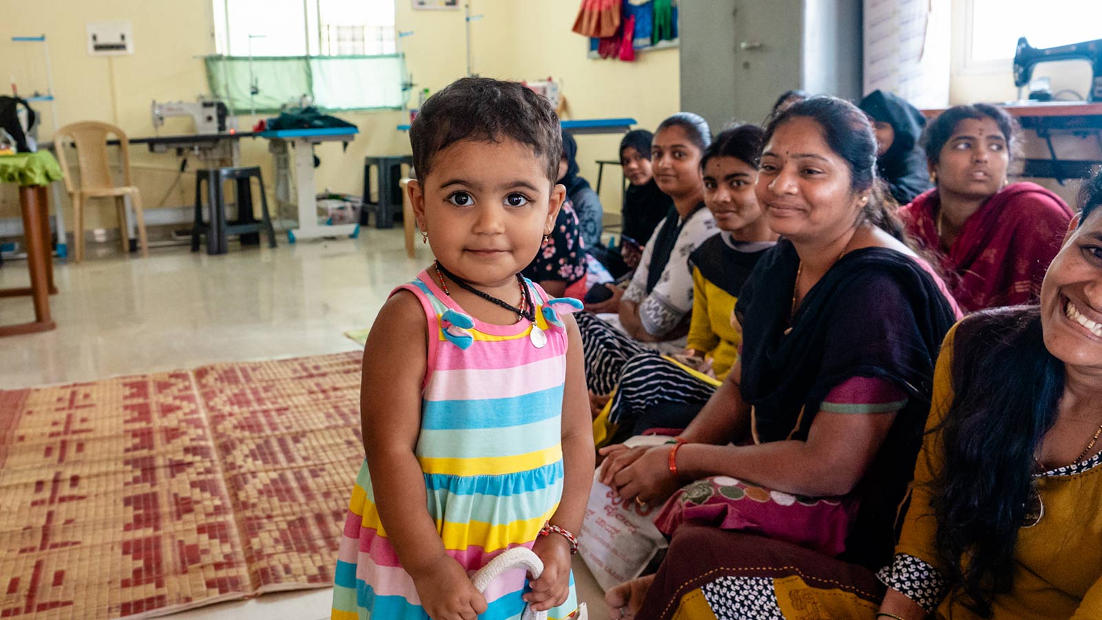 a little girl in a striped dress smiles shyly with a group of women sitting in the background at a women's center in India