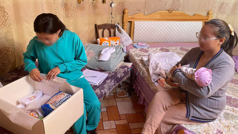 Single mother in turquoise-colored sweatsuit looks at items in baby box, while another single mother holds a baby in pink hat