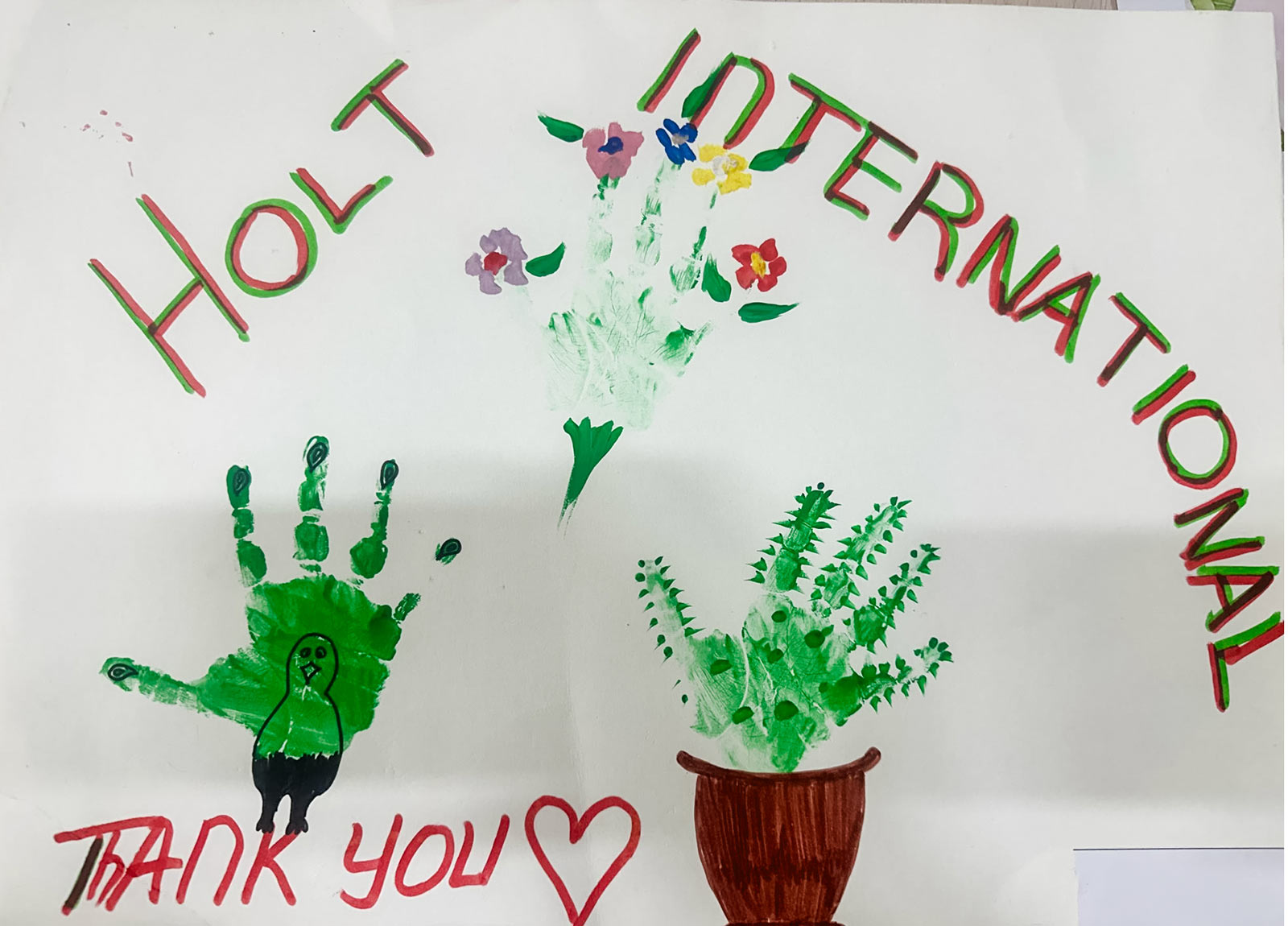 three green painted handprints of a sponsored child create the shape of a turkey, a bouquet and a potted cactus with the words "Holt International Thank You"