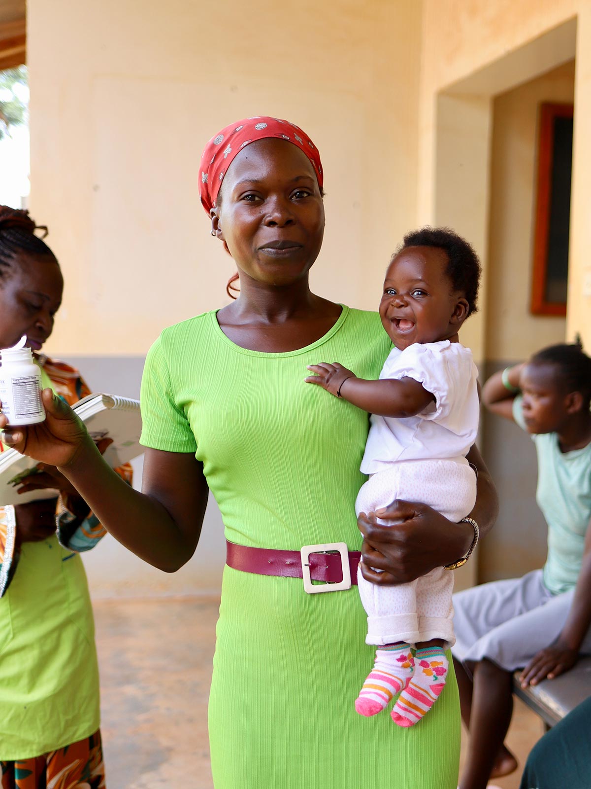 A mother in a green dress holds her baby in one arm and a bottle of vitamins in the other during a health event in Uganda
