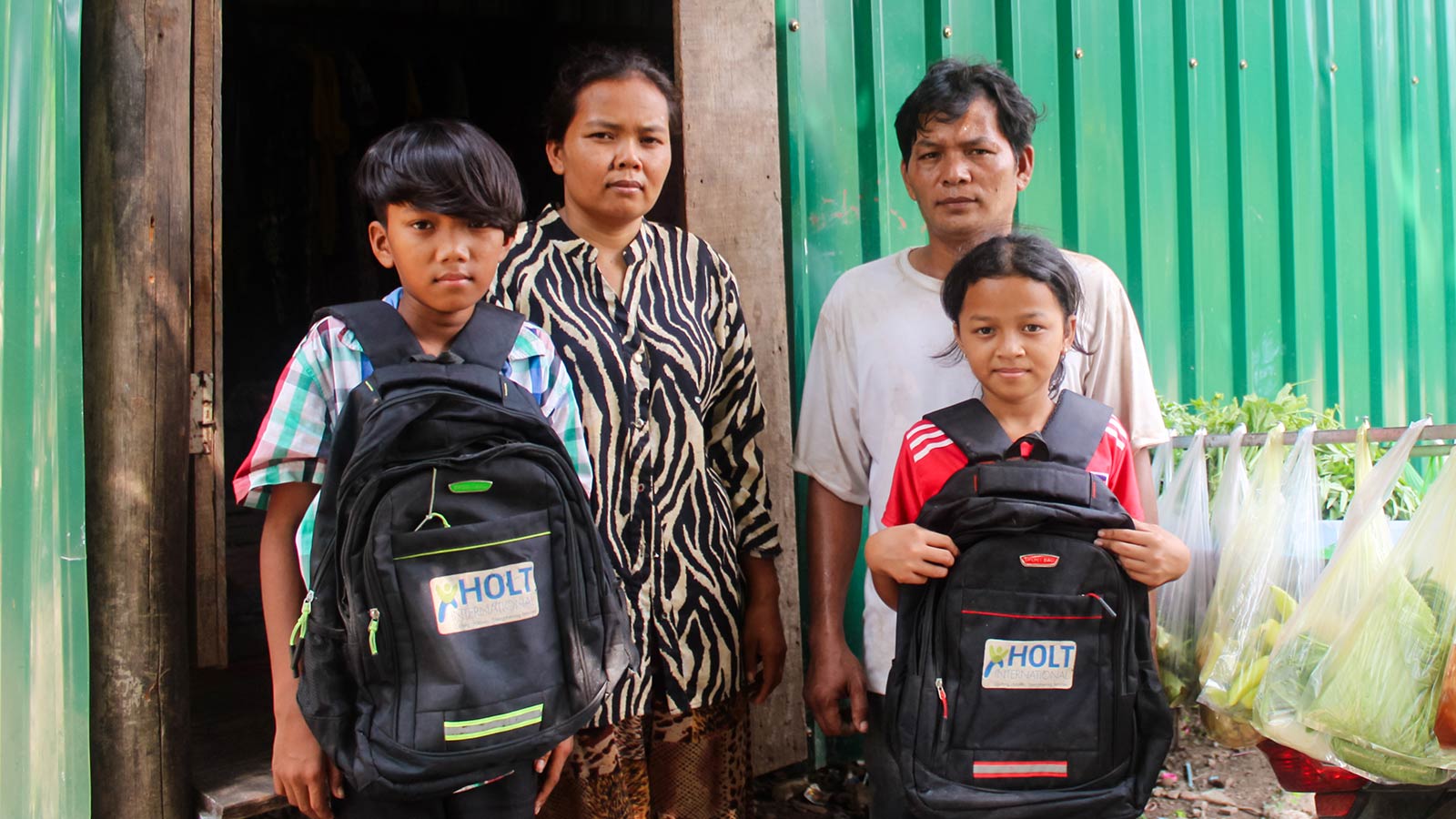 sponsored twins in Cambodia show off their school backpacks with their parents in the background