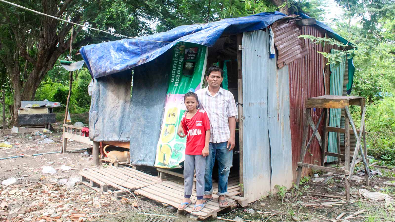 A father and daughter stand outside their makeshift house, made with tin sheets and tarps