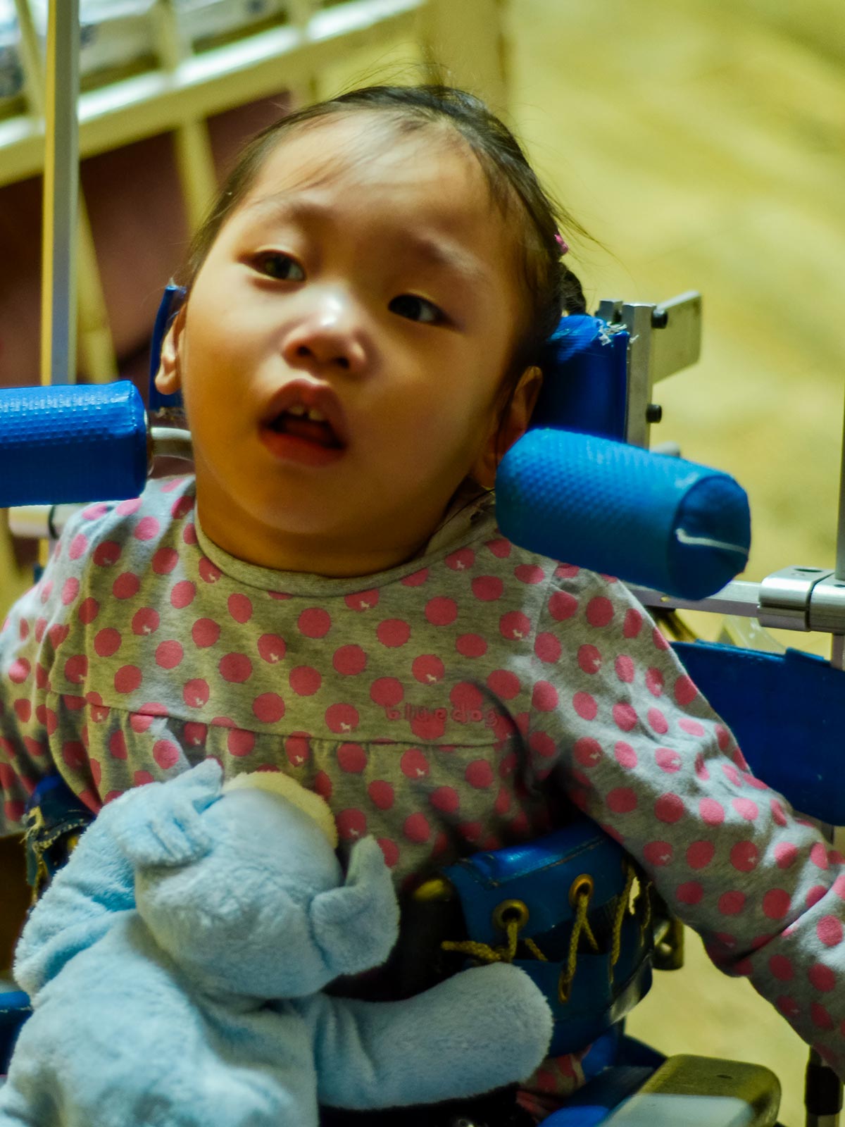a girl sits in a supportive chair with a stuffed animal on her lap as she receives nutrition through a feeding tube at Ilsan in Korea