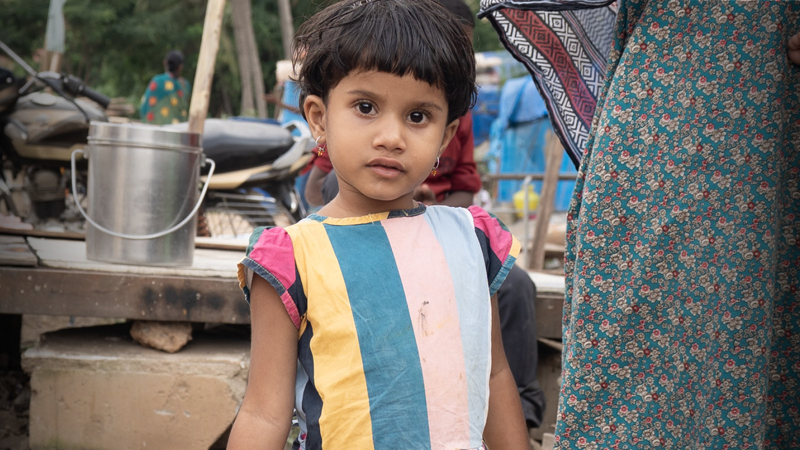 Girl in striped dress looking at camera. Trash and debris behind her.