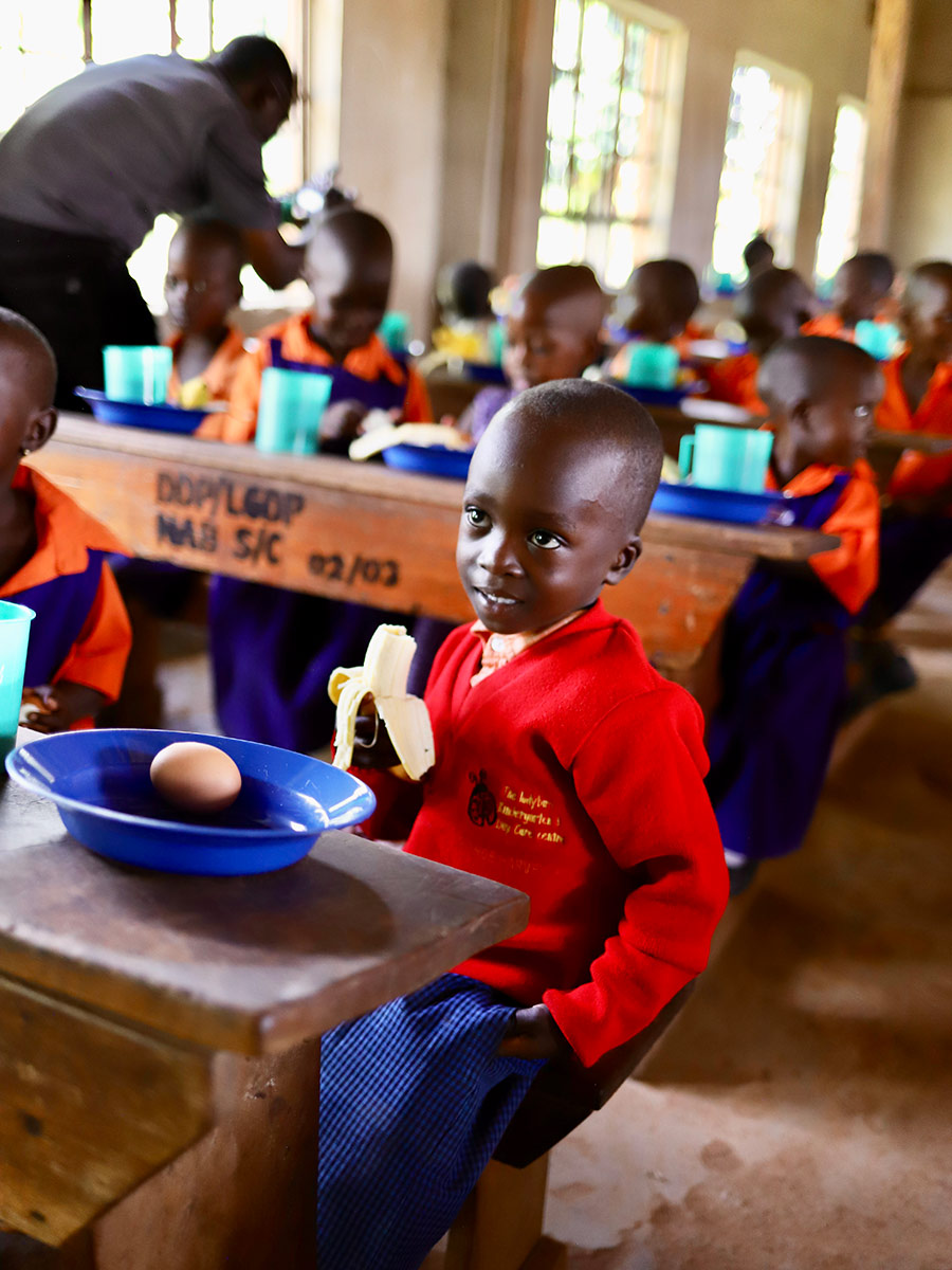 a boy in a school uniform holds a banana and smiles with his school lunch in Uganda