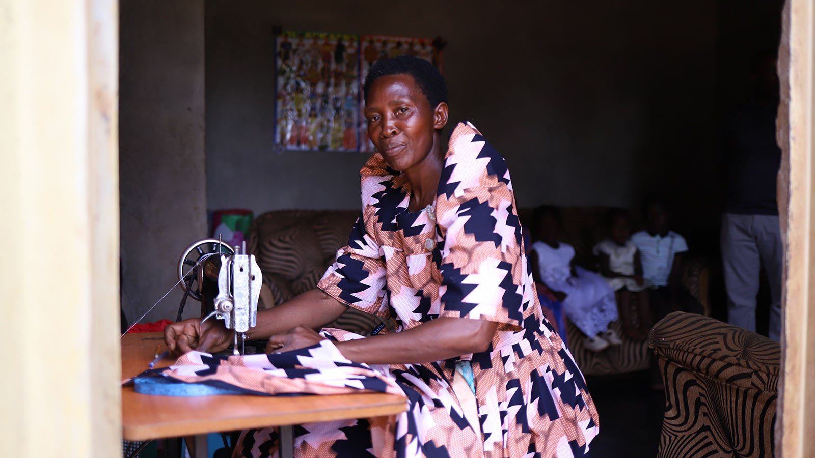 a woman in a patterned pink and black dress sews with a sewing machine inside her home in Uganda
