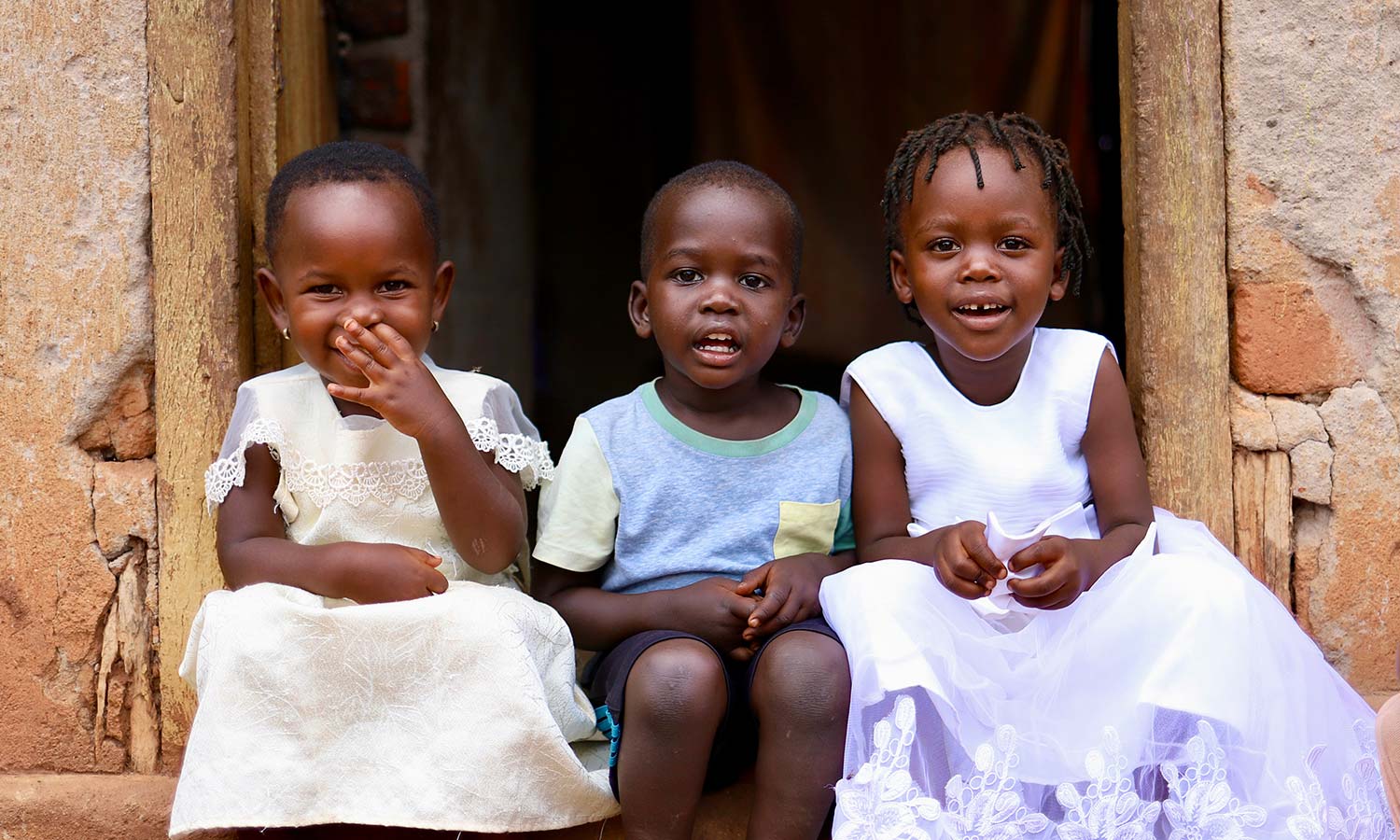 two little girls and a boy smile shyly on the doorstep of their home in rural Uganda