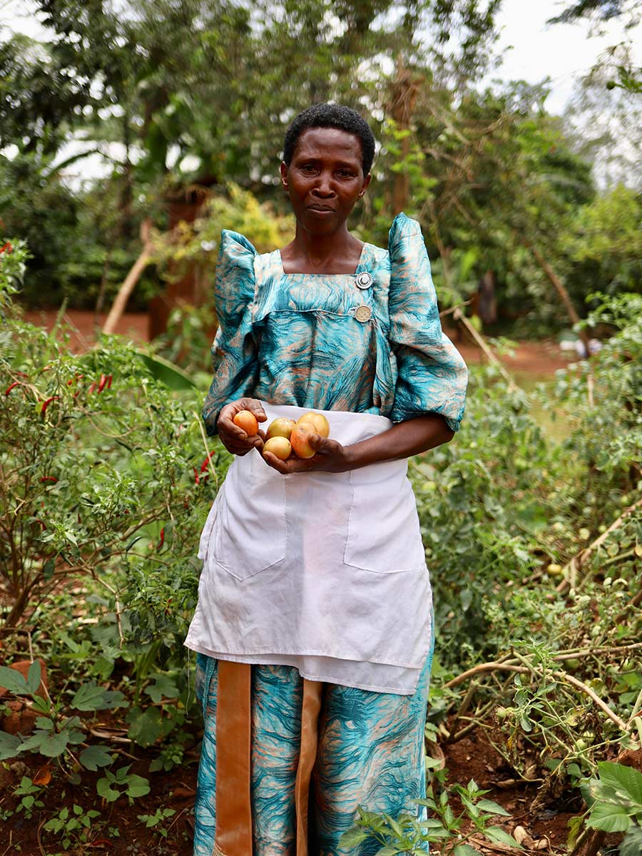 a woman wearing a teal dress and an apron holds vegetables she grew in her garden in Uganda