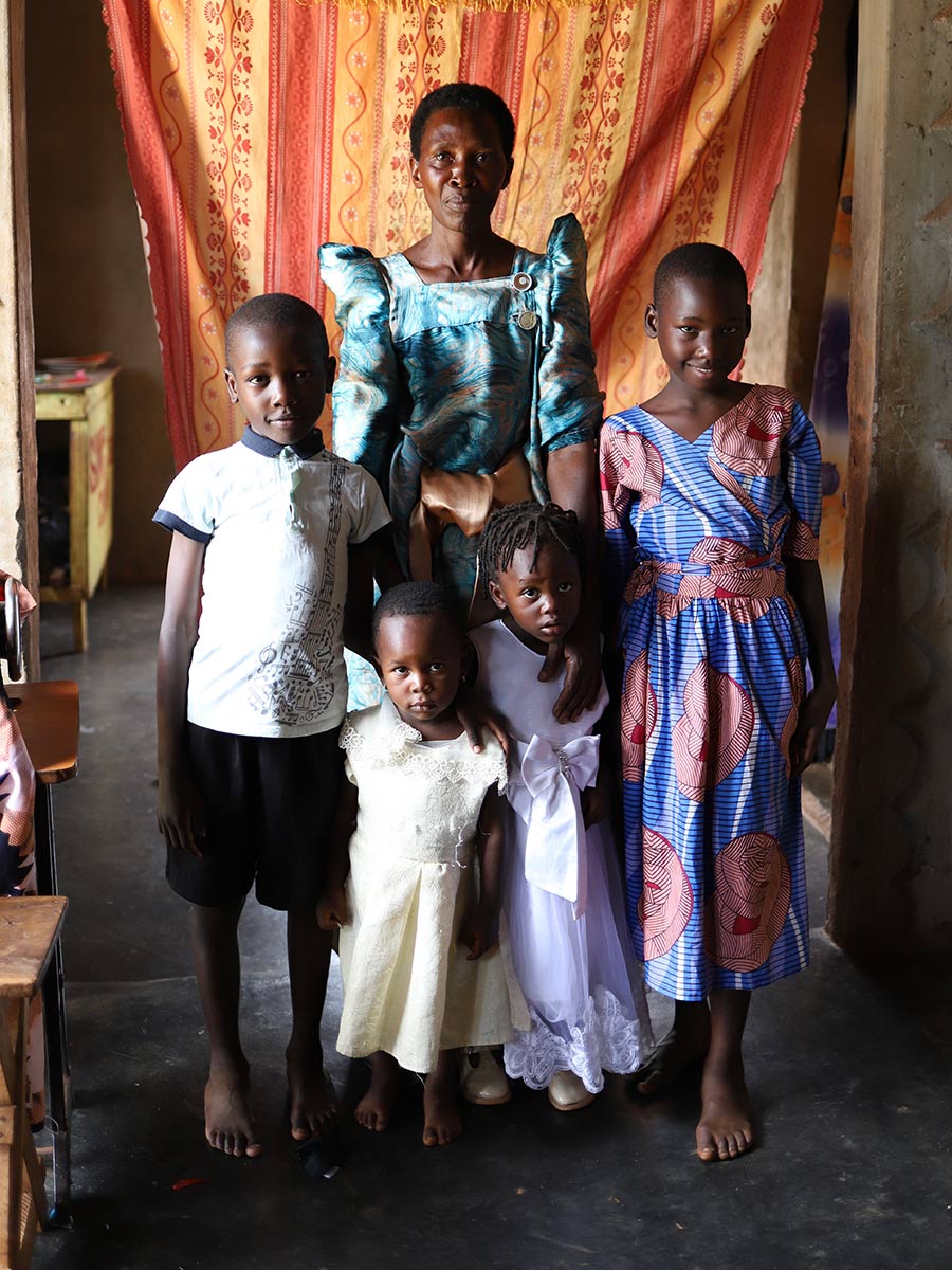 a woman in a teal dress stands with her four grandchildren at home in rural Uganda