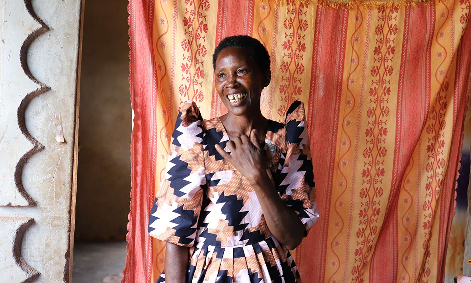 a woman in a patterned pink and black dress smiles inside her home in Uganda